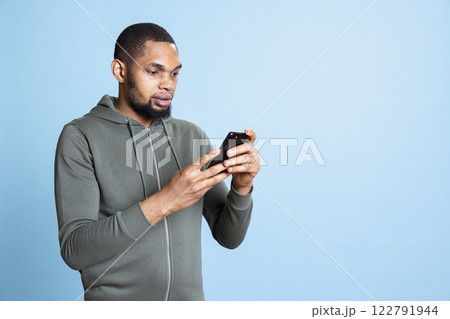 Optimistic young adult answering text messages on smartphone, browsing online websites and surfing the web. Lively guy using social media applications against blue background in studio. 122791944