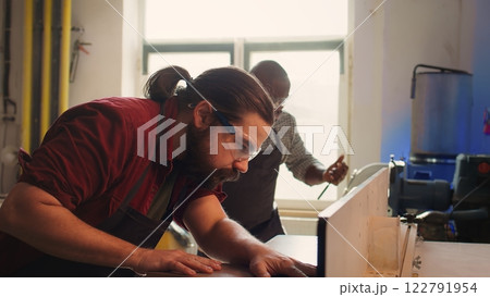 Woodworking expert wears protection gear, inserting plank in spindle moulder next to coworker. CNC machinist uses safety glasses while cutting lumber with wood shaper helped by colleague, camera B 122791954