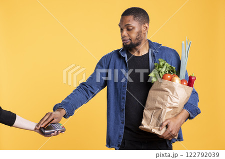 Young adult paying with a card at the pos terminal in studio, purchasing a bag of fresh homegrown produce in a paper bag. Person using electronic banking payment during shopping. Young adult paying with a card at the pos terminal in studio, purchasing a bag of fresh homegrown produce in a paper bag. Person using electronic banking payment during shopping. 122792039