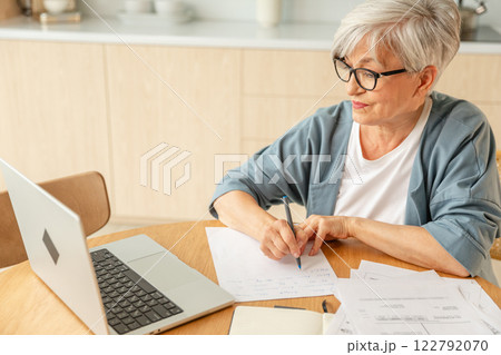 Middle aged senior woman using laptop computer writing notes at home. Focused mature old businesswoman work in home office making written records doing online work. Mature boss professional worker 122792070