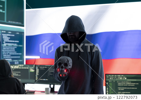 Conceptual image of a hooded russian hacker creating hazardous alerts, holds a skull and warning of death threats. Russian federation works on cybercrime and violence, panic and torment. 122792073
