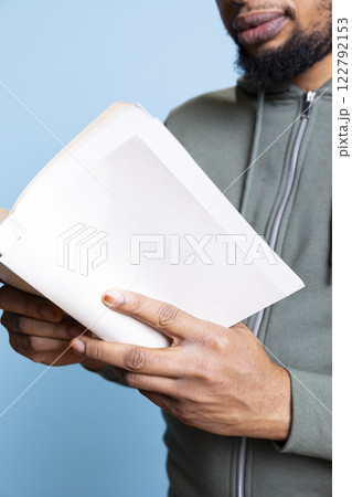 African american cheerful guy enjoys reading a book in studio, feeling happy with literature and novel against blue background. Confident book lover analyzing the synopsis and characters. Close up. African american cheerful guy enjoys reading a book in studio, feeling happy with literature and novel against blue background. Confident book lover analyzing the synopsis and characters. Close up. 122792153