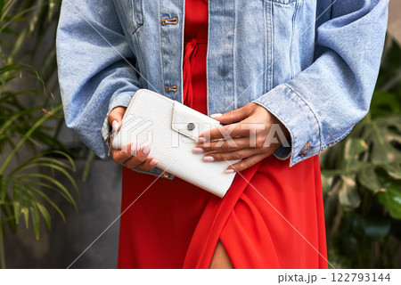 Elegant woman in denim jacket and red dress holding stylish white wallet outdoors, fashion and lifestyle concept 122793144