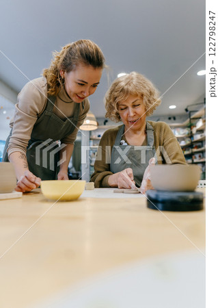 Two women work together with clay, developing skills and enjoying the artistic process. Elderly Caucasian woman learns pottery at a creative workshop with a young mentor 122798247