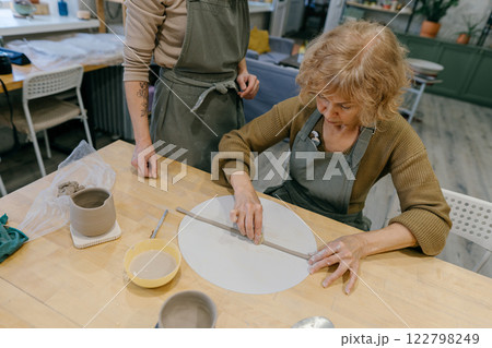 Teacher guides an elderly women, demonstrating sculpting techniques in a hands-on class. Elderly Caucasian woman learns pottery at a creative workshop with a young mentor 122798249