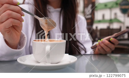 A person leisurely stirring a delicious hot beverage while holding their phone in a cozy outdoor ambiance 122799075