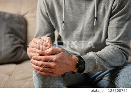 Man holding knee in pain while sitting on couch Man holding knee in pain while sitting on couch 122800725