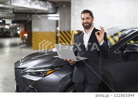 Caucasian male businessman in suit holding laptop, showing OK sign in parking garage. Represents success, confidence, and modern technology. Scene conveys business and automotive themes. Caucasian male businessman in suit holding laptop, showing OK sign in parking garage. Represents success, confidence, and modern technology. Scene conveys business and automotive themes. 122801044