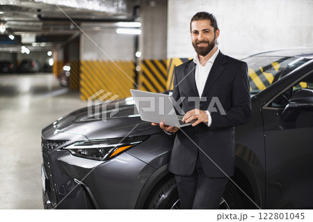 Confident Caucasian businessman stands with laptop next to car in underground garage. Man wearing suit works remotely, showcasing modern technology and urban lifestyle. Confident Caucasian businessman stands with laptop next to car in underground garage. Man wearing suit works remotely, showcasing modern technology and urban lifestyle. 122801045
