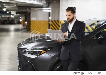Caucasian male businessman using laptop in parking garage beside modern vehicle. Professional setting, focused on work, wearing stylish suit 122801048