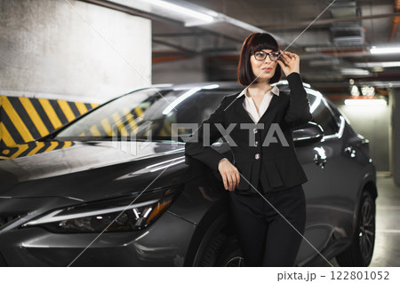 Young Caucasian woman in professional attire stands confidently by car in garage. Emphasizes leadership and style. Business setting with indoor parking background. Young Caucasian woman in professional attire stands confidently by car in garage. Emphasizes leadership and style. Business setting with indoor parking background. 122801052