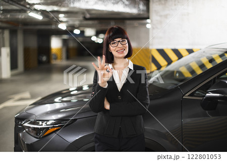 Young Caucasian woman in business attire standing near car in garage making okay sign. Represents confidence, success, approval. Suitable for business, automotive, professional themes. Young Caucasian woman in business attire standing near car in garage making okay sign. Represents confidence, success, approval. Suitable for business, automotive, professional themes. 122801053
