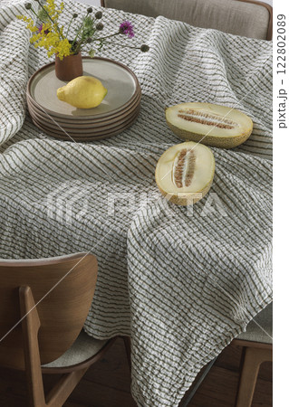 Simple organisation of dining table with ceramic dinnerware, linen tablecloth, dining chairs, half sliced melon, lemon and wild spring flowers. Home decoration concept 122802089