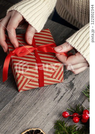 Female hands in a white sweater sleeves tying a bow on a Christmas gift box on a wooden background, vertical image, close-up. Female hands in a white sweater sleeves tying a bow on a Christmas gift box on a wooden background, vertical image, close-up. 122802344