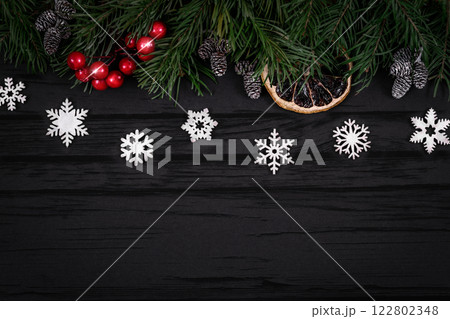 Christmas decorative upper border with green spruce, orange slice, cones, white snowflakes and holly berries on black wooden background, top view, copy space. Christmas decorative upper border with green spruce, orange slice, cones, white snowflakes and holly berries on black wooden background, top view, copy space. 122802348