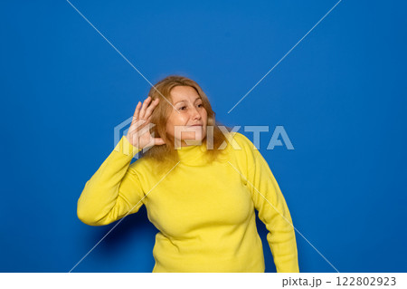 Caucasian woman in her 40s wearing a yellow sweater with one hand on her ear eavesdropping on something, isolated over blue background. Gossip and rumors concept Caucasian woman in her 40s wearing a yellow sweater with one hand on her ear eavesdropping on something, isolated over blue background. Gossip and rumors concept 122802923
