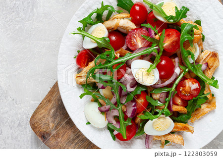 a warm salad with fried chicken fillet, arugula and cherry tomatoes, and boiled quail eggs, on a white plate, no people. 122803059