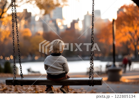 A toddler sits alone on a swing, gazing at a cityscape blurred in the autumnal sunset, conveying a sense of quiet contemplation 122805134