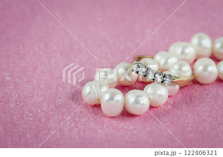 A gold ring with diamonds dressed on a pearl necklace shot against a pink background. 122806321