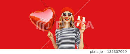 Happy laughing young woman holding red heart shaped balloon, gift box isolated on studio background 122807069