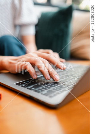 Hands typing on a laptop keyboard in a cozy home environment with natural lighting and a wooden surface. 122808326