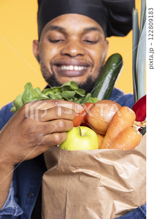 African american chef admiring organic produce in a paper bag, smelling the fresh aroma of fruits and vegetables in studio. Person cooking with bio natural ingredients. Close up. African american chef admiring organic produce in a paper bag, smelling the fresh aroma of fruits and vegetables in studio. Person cooking with bio natural ingredients. Close up. 122809183