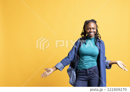 Cheerful african american woman smiling, doing shrugging shoulders gesture. Easygoing jolly BIPOC teenager doing nonchalant carefree hand gesturing, isolated over studio background 122809270
