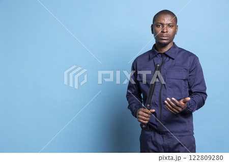 Security agent holding his baton weapon while being on duty, working on ensuring protection and safety. African american armed bodyguard with authority poses against blue background. Security agent holding his baton weapon while being on duty, working on ensuring protection and safety. African american armed bodyguard with authority poses against blue background. 122809280