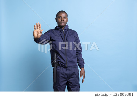 African american security warden does a stop sign with his palm raised and saying no, standing against blue background. Professional safeguarding officer does not let people pass, authority. African american security warden does a stop sign with his palm raised and saying no, standing against blue background. Professional safeguarding officer does not let people pass, authority. 122809391
