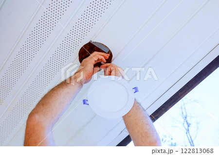 Hands are fixing an LED ceiling light fixture in brightly lit soffit with white ceiling. 122813068
