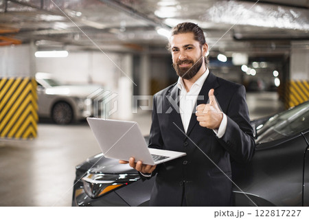 Caucasian male businessman in suit holding laptop with thumbs up gesture in underground parking lot. Professional and confident, conveying success and positivity. 122817227