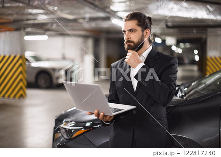 Caucasian male businessman in suit standing in parking garage using laptop, appearing thoughtful and focused. Middle-aged professional contemplating work decisions in modern business environment. 122817230