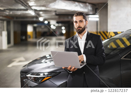 Professional Caucasian businessman in suit works on laptop while standing in underground parking garage. Focus on business, technology, multitasking, urban setting. 122817237