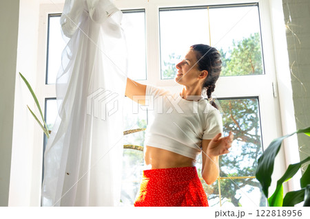 Woman hangs transparent tulle curtains on large windows in the house inside the interior with potted plants. Spring cleaning, tidying up Woman hangs transparent tulle curtains on large windows in the house inside the interior with potted plants. Spring cleaning, tidying up 122818956
