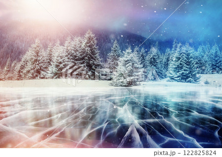 Blue ice and cracks on the surface of the ice. Frozen lake in winter mountains. It is snowing. The hills of pines. Winter. Carpathian Ukraine Europe 122825824