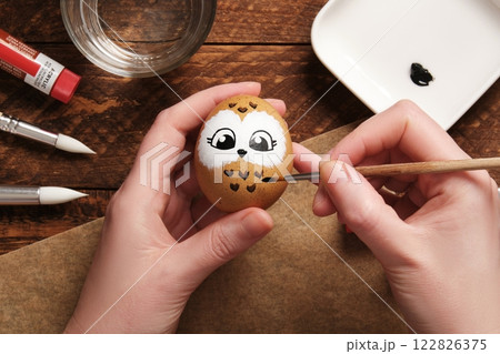 Hands drawing the face of an owl on a brown eggshell. Preparation for the celebration of Easter. Painting Easter eggs with acrylic pigment dyes in tubes. 122826375