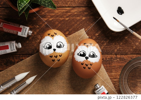 Easter eggs with owls, hand-painted with acrylic paints, on a background of paints, brushes, and palette. DIY concept 122826377