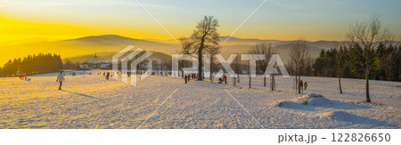 At sunset in Prichovice, Czechia, people enjoy the winter landscape covered in snow. The vibrant colors of the sky create a stunning contrast with the white terrain and distant hills. At sunset in Prichovice, Czechia, people enjoy the winter landscape covered in snow. The vibrant colors of the sky create a stunning contrast with the white terrain and distant hills. 122826650