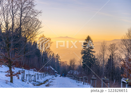 A winter sunset bathes the snow-covered road in Prichovice, Czechia. In the background, Jested Mountain stands majestically against the glowing horizon. 122826651
