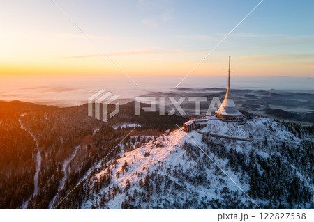 Stunning view of Jested Mountain at sunrise during winter. The sun casts a warm glow over the snow-covered slopes and the unique tower stands tall against the clear sky. 122827538