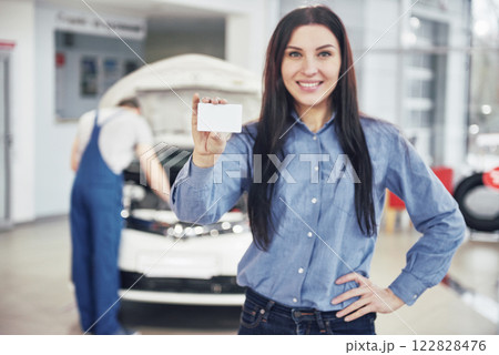 A beautiful woman keeps a business card of the car service center. The mechanic inspects the car under the hood in the background 122828476