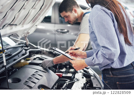 A man mechanic and woman customer look at the car hood and discuss repairs A man mechanic and woman customer look at the car hood and discuss repairs 122828509