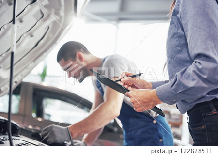 A man mechanic and woman customer look at the car hood and discuss repairs A man mechanic and woman customer look at the car hood and discuss repairs 122828511