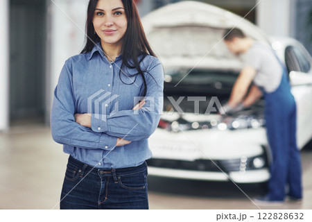 Woman at a car garage getting mechanical service. The mechanic works under the hood of the car 122828632