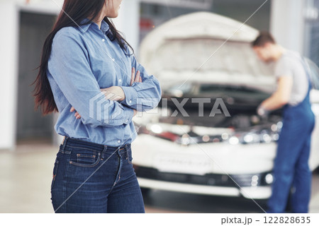 Woman at a car garage getting mechanical service. The mechanic works under the hood of the car 122828635