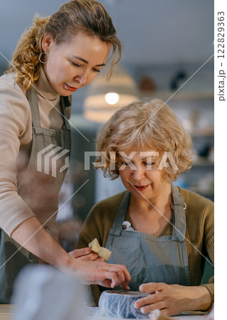 Teacher guides an elderly student, demonstrating sculpting techniques in a hands-on class. Senior woman practices clay modeling with help from a younger instructor in a ceramics studio. 122829363