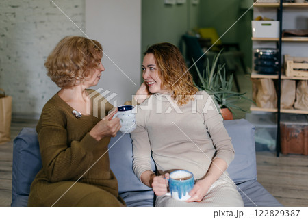 Mother and daughter, two Caucasian women of different ages, sit together on a couch with tea. The elderly mother and young daughter smile while enjoying a warm and relaxed conversation 122829387