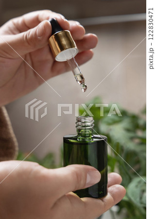 Closeup of woman hands opening green dropper bottle aginst green plant, drop falling from pipette Closeup of woman hands opening green dropper bottle aginst green plant, drop falling from pipette 122830471
