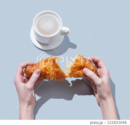 Hands breaking a fresh croissant over a blue background with a cup of cappuccino or latte coffee. 122831048