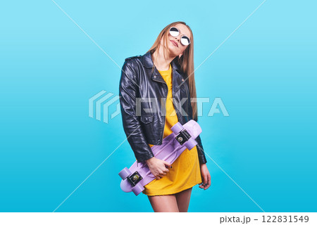 Attractive tanned girl with smile in face wears black leather jacket posing with pleasure. Indoor photo of lovely woman in sunglasses standing with skateboard on blue background Attractive tanned girl with smile in face wears black leather jacket posing with pleasure. Indoor photo of lovely woman in sunglasses standing with skateboard on blue background 122831549